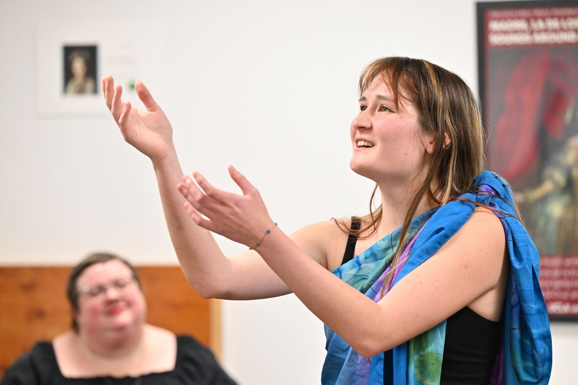 Person in a colorful scarf gestures animatedly while speaking. Another person watches in the background.