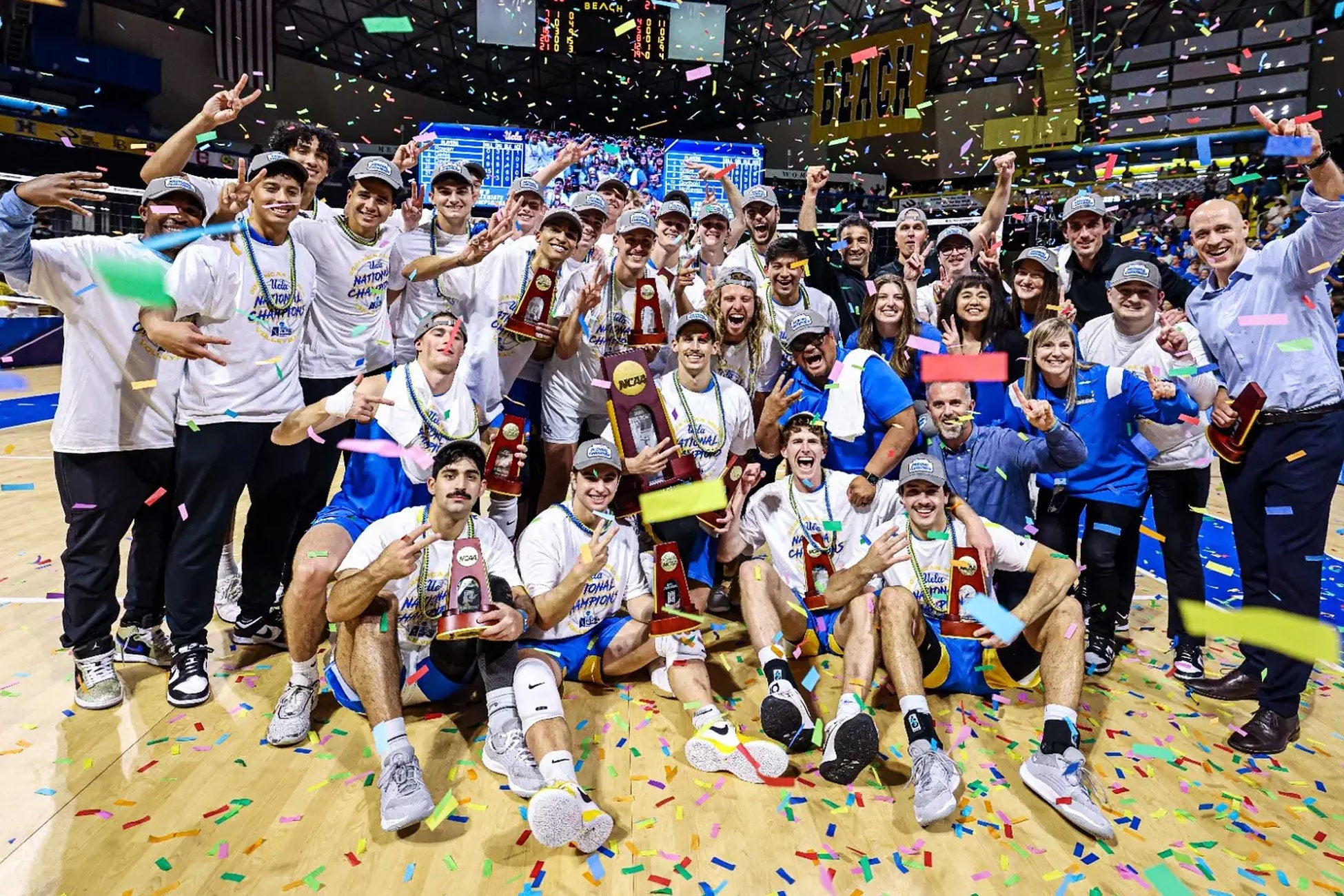 Celebratory photo of the UCLA men&#039;s volleyball team and coaches featuring flying multi-colored confetti, smiling faces, and a large NCAA trophy. 