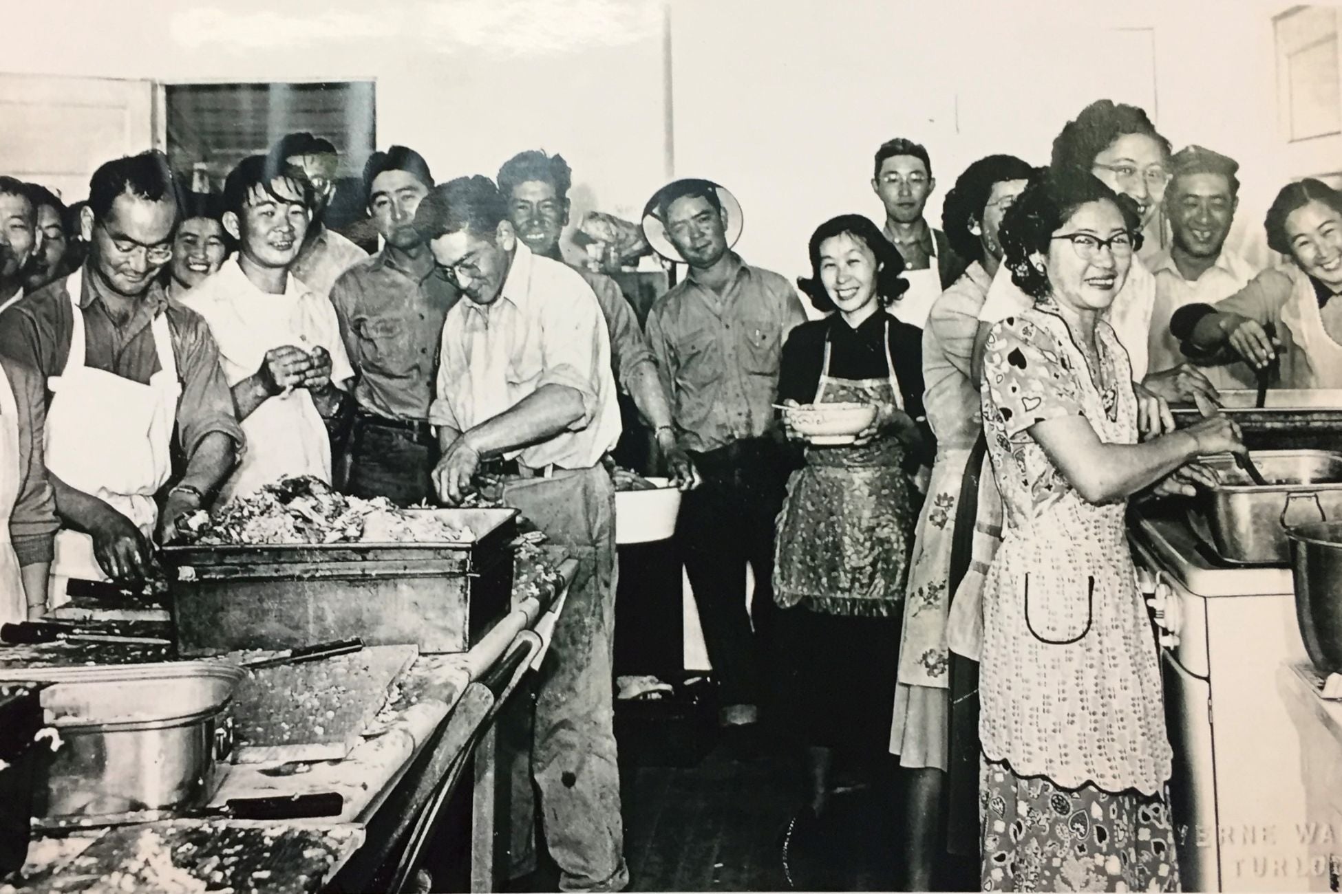 Black-and-white image of about 20 Japanese American men and women preparing meal in shared kitchen