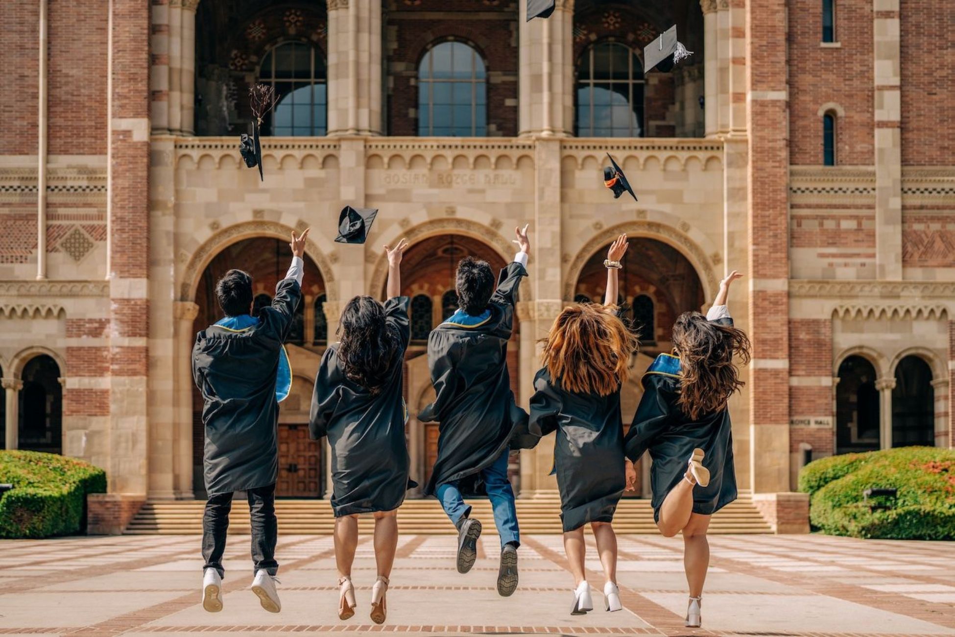 Five graduating students dressed in black gowns, throwing their caps in the air in front of Royce Hall