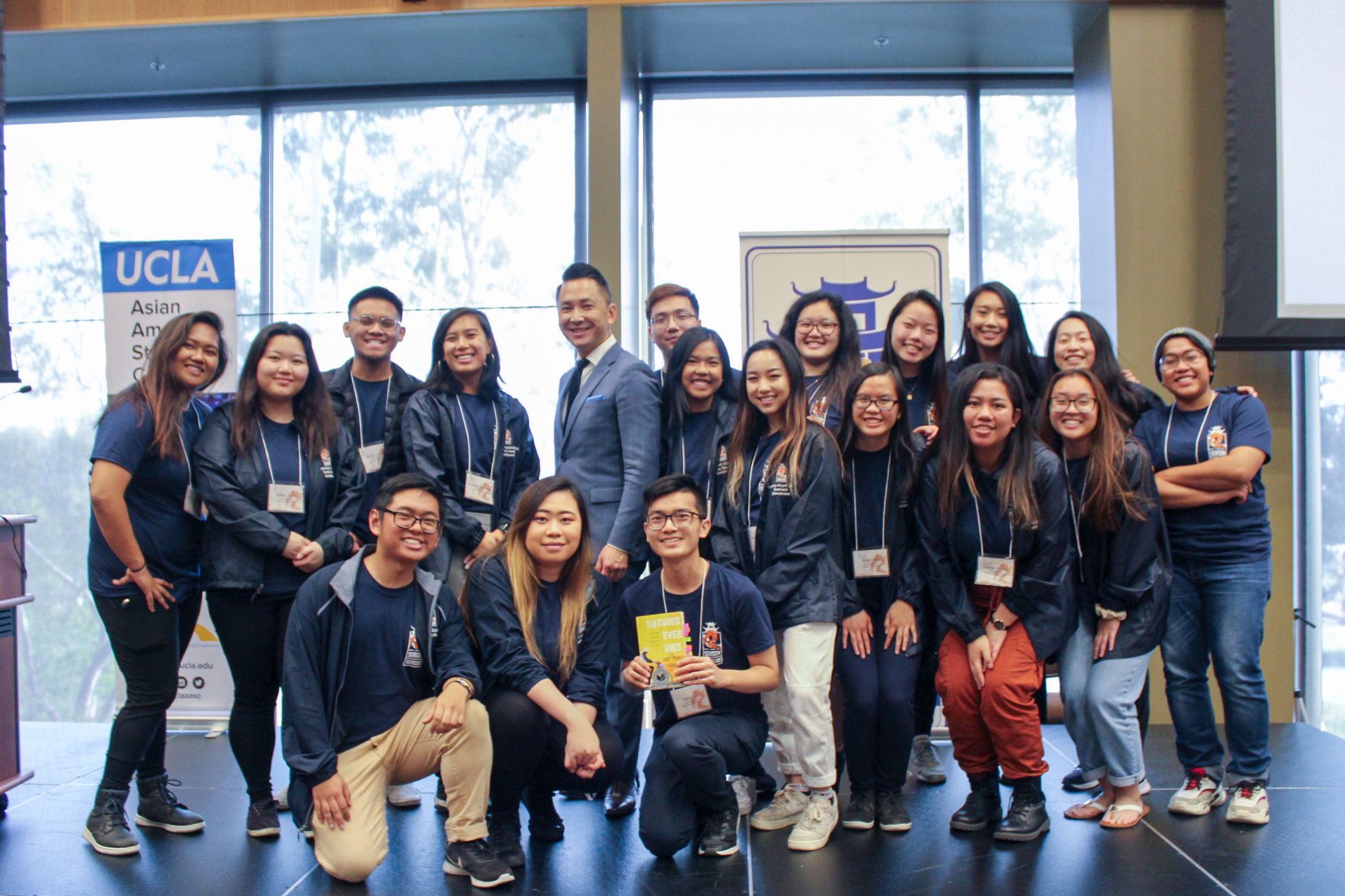 Group of Southeast Asian students and a professor smiling as they stand and kneel together on stage at a conference