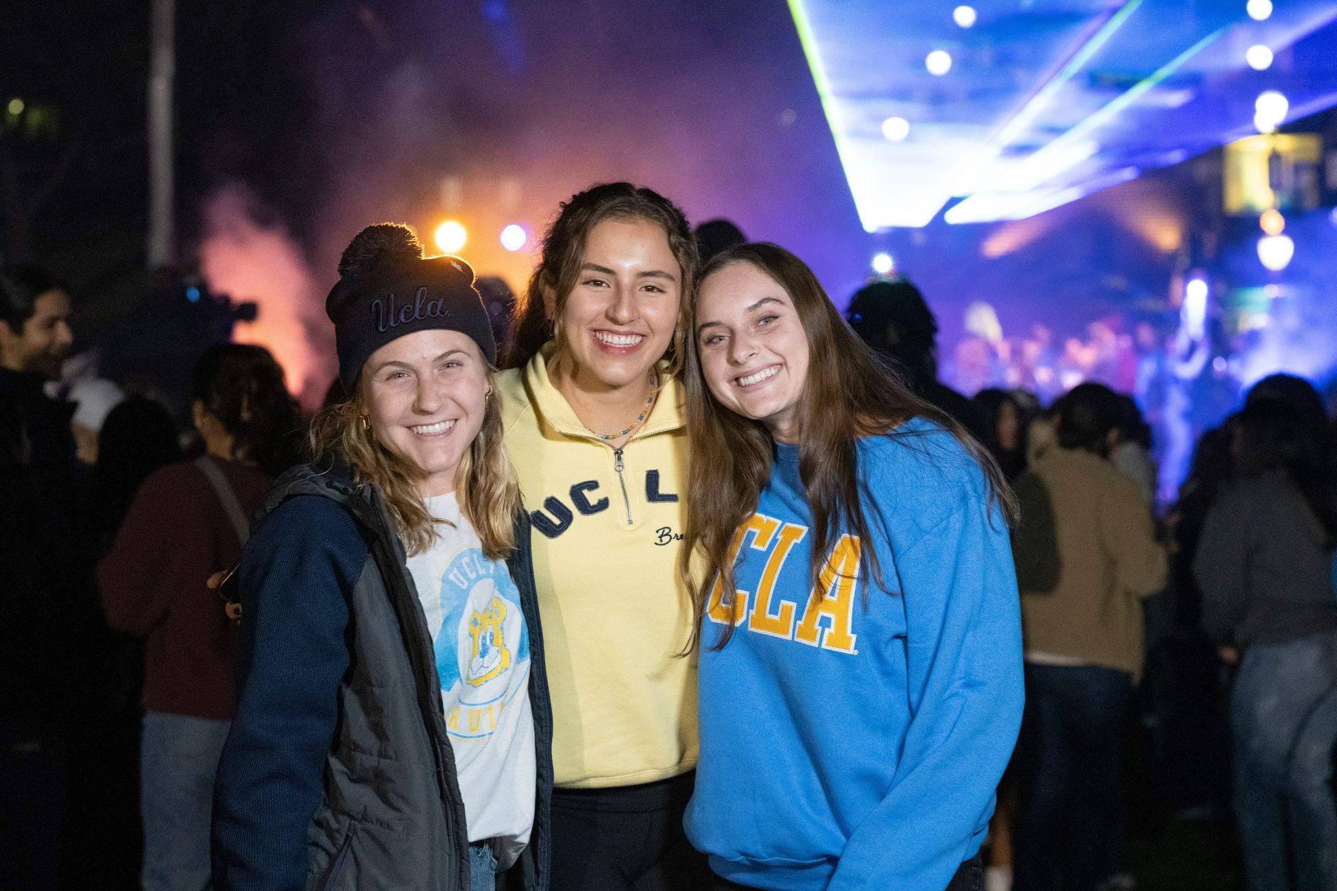 Three students smiling and posing together at a lively concert,  with a backdrop of colorful lights and an energetic crowd