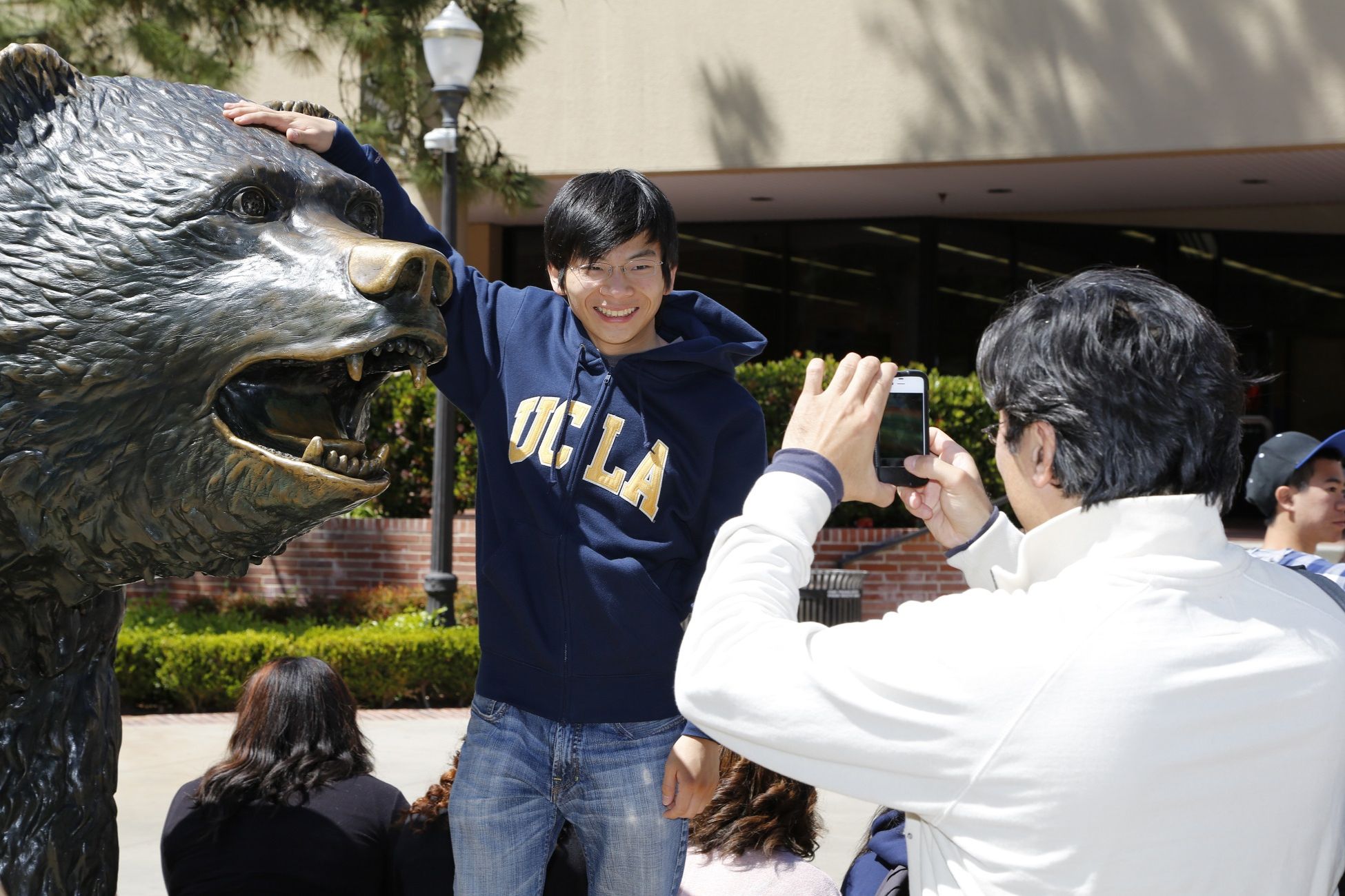 Young man in blue UCLA hoodie places right hand on Bruin statue&#039;s head, smiles at man taking photo