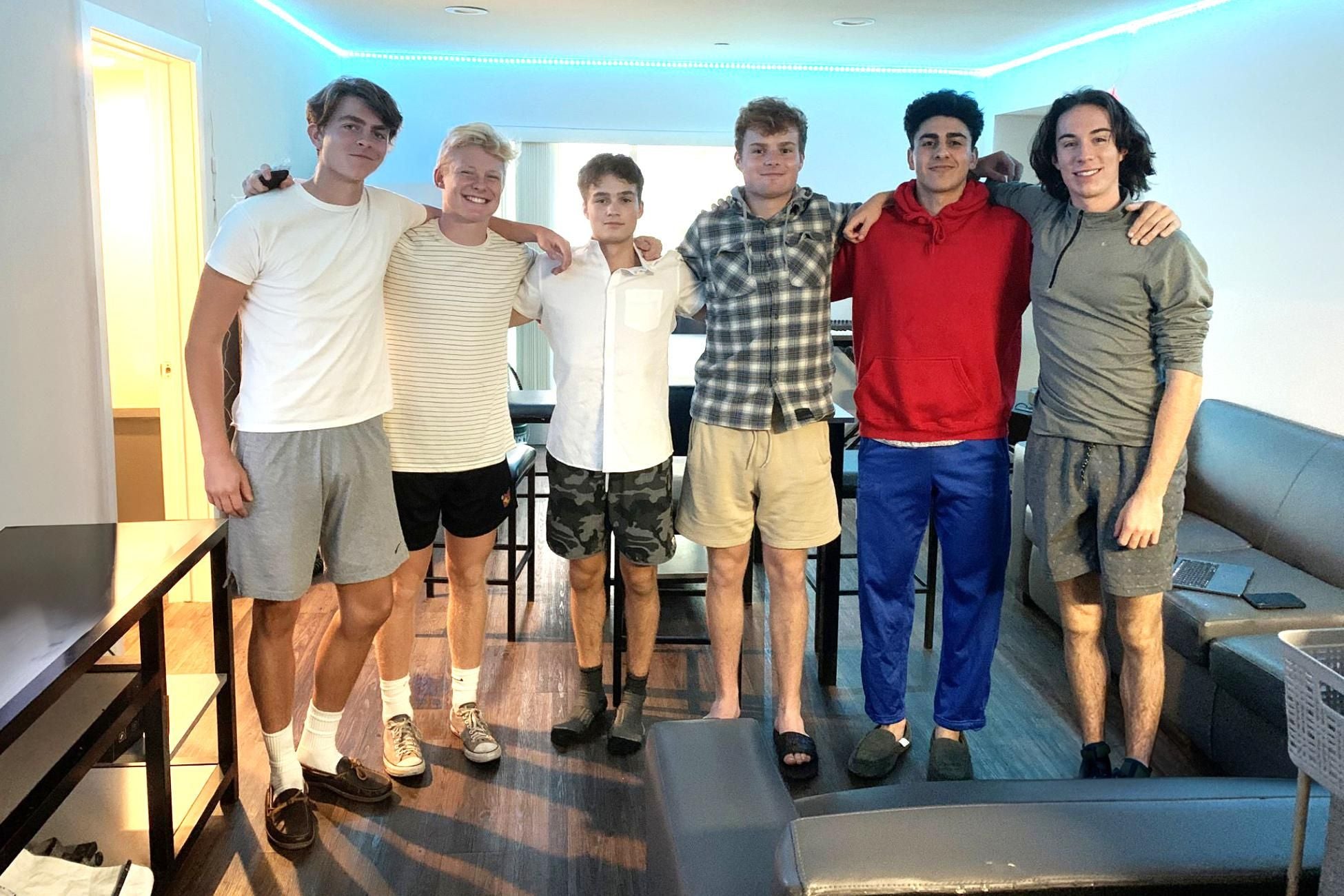 Six young men stand with their arms around each other&#039;s shoulders and smile to camera