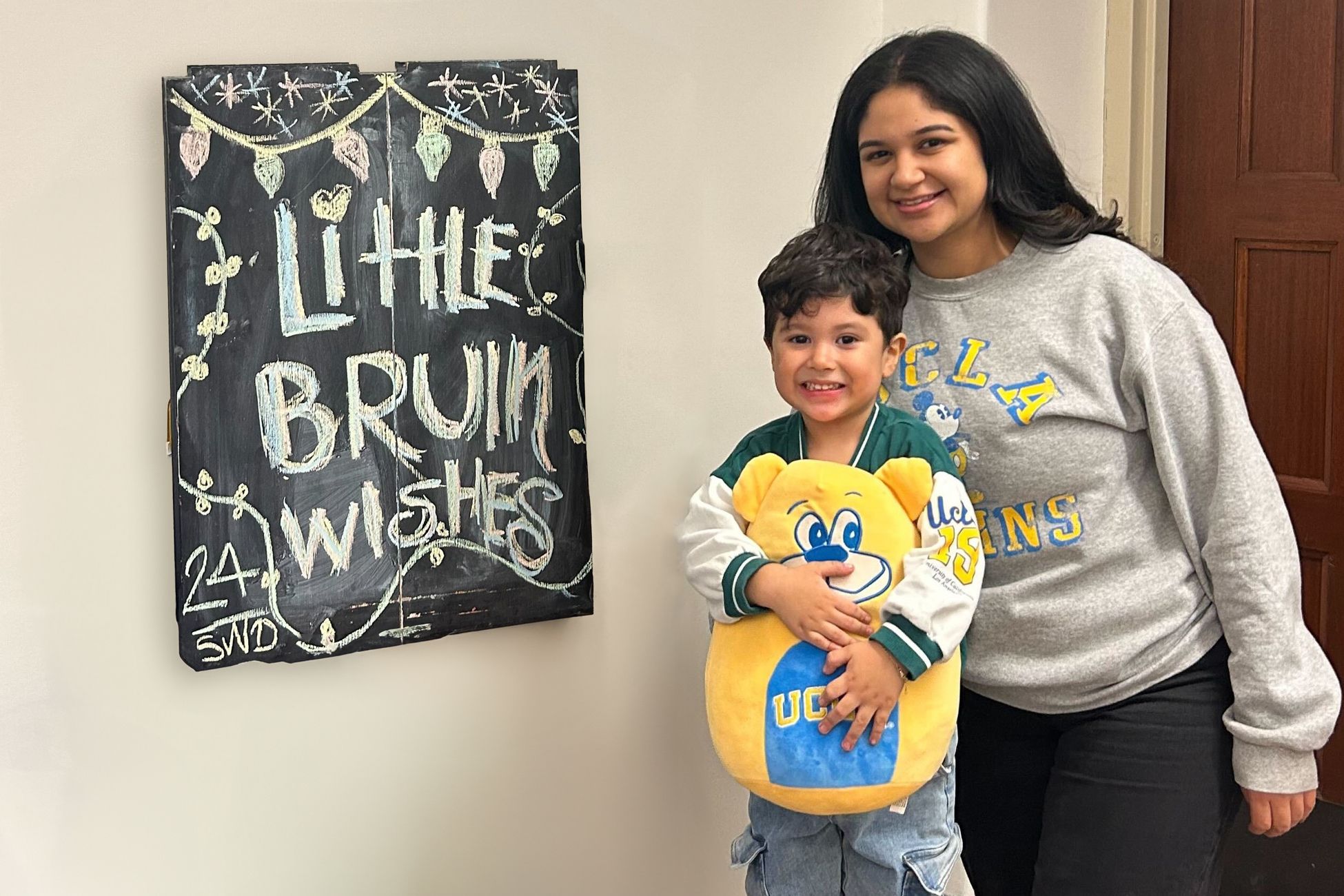 Chalkboard reads &quot;Little Bruin Wishes&quot; next to boy holding Joe Bruin plush, standing next to woman