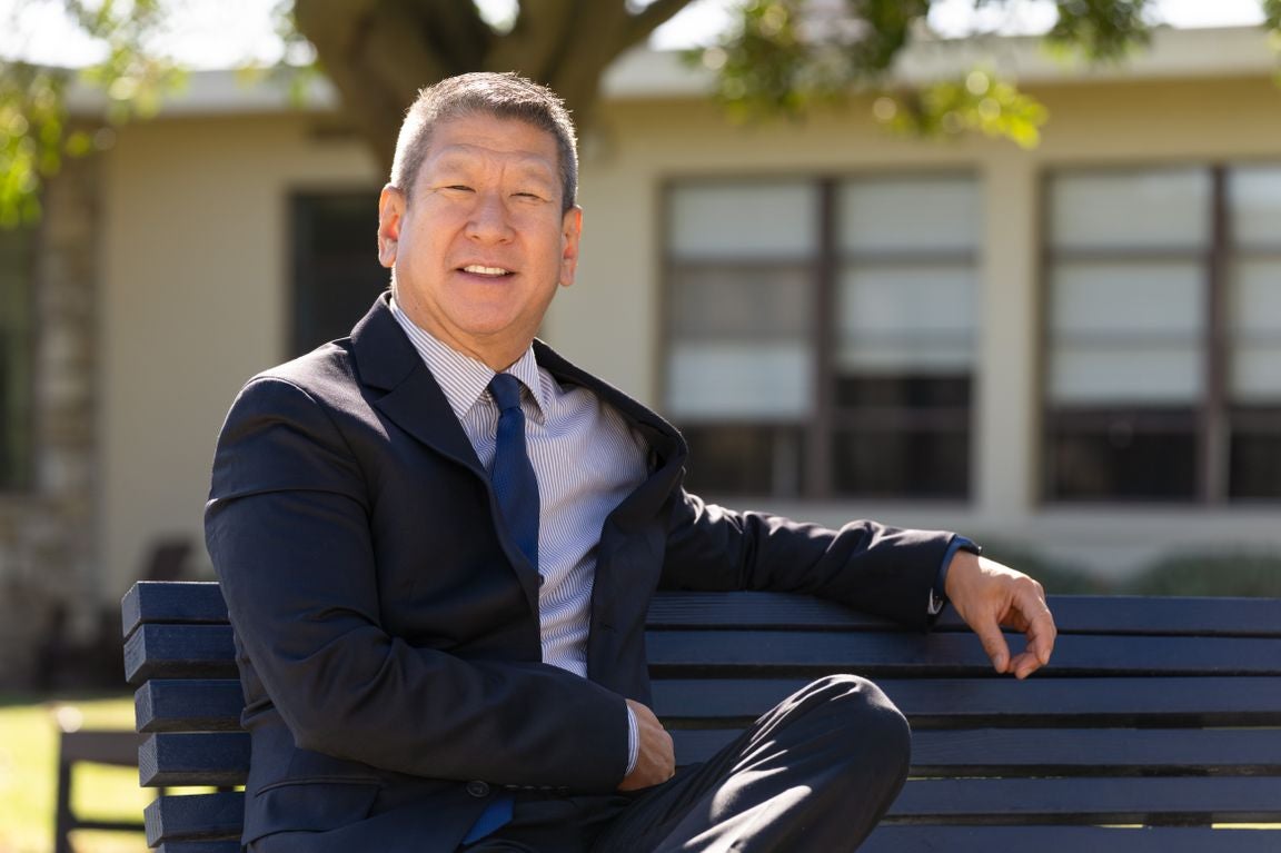 Steve Yu, wearing a suit and tie, sits on a bench, posing and smiling