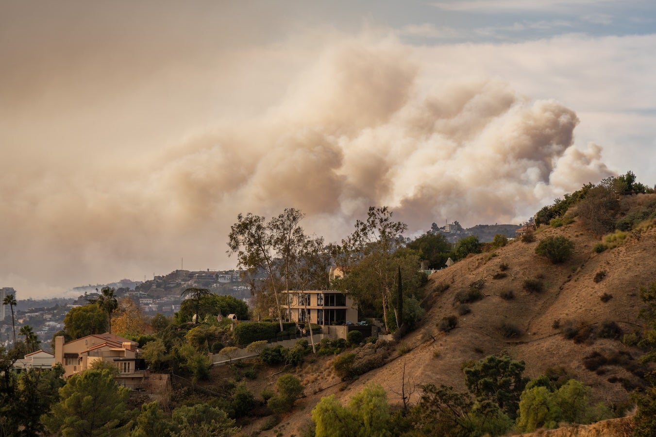 Large wildfire smoke cloud rising behind a hill with houses 