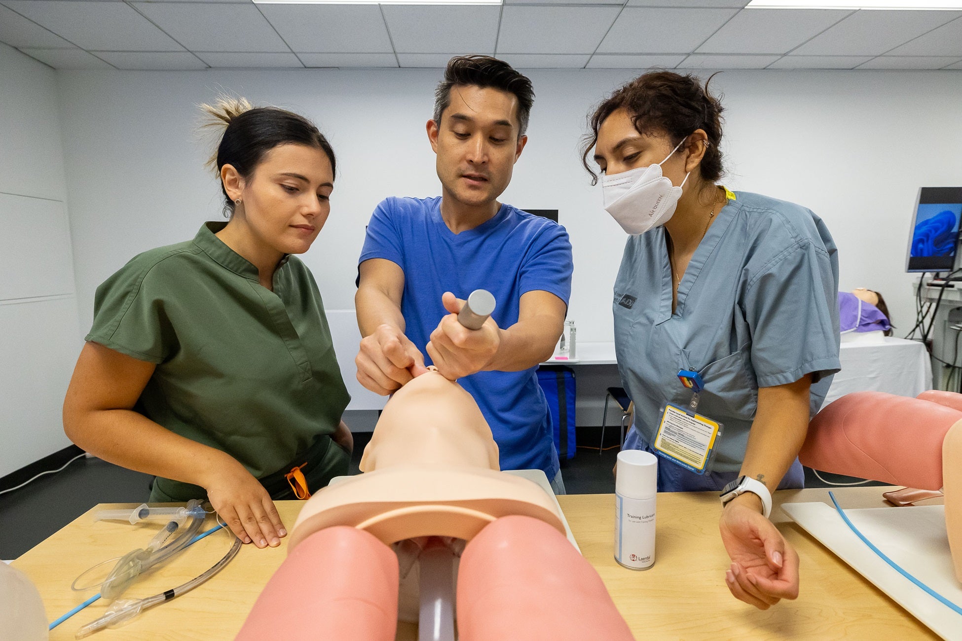 Dark-haired man demonstrates using a scope on a manikin to two women in scrubs, one wearing a mask