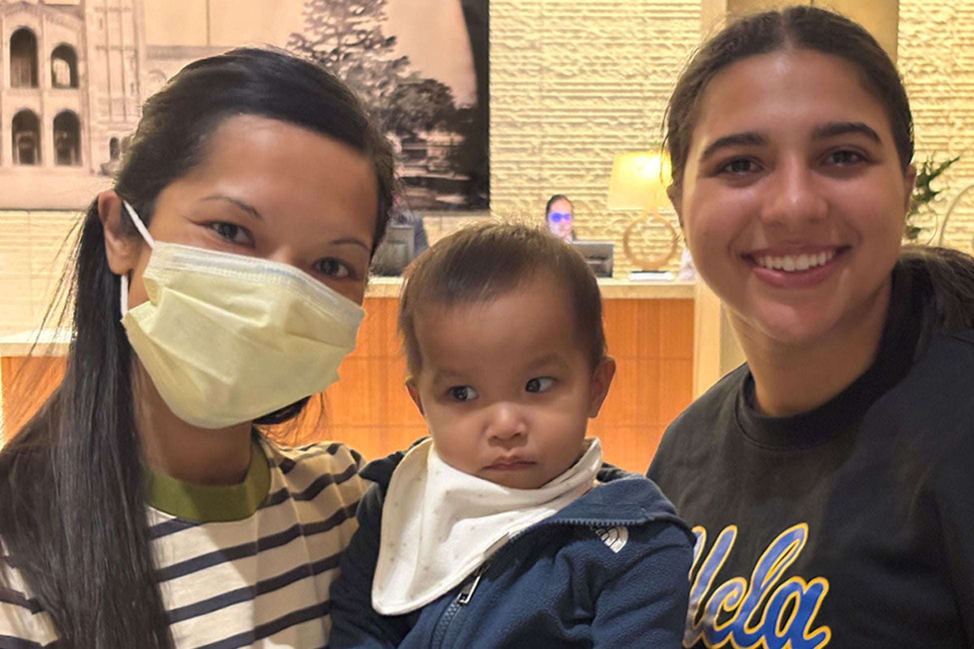 Two women, one wearing a mask and the other smiling, pose with an infant