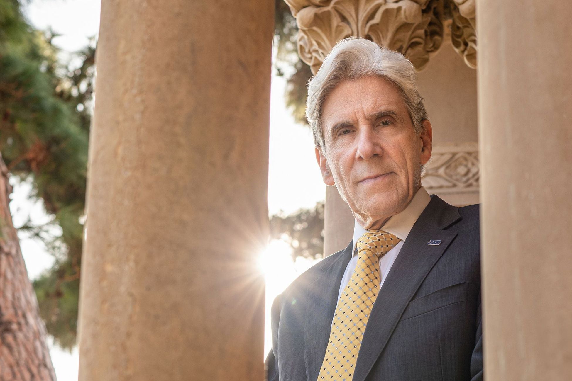 Chancellor Frenk in a suit and tie stands in front of grand pillars with the sun peeking behind his shoulder