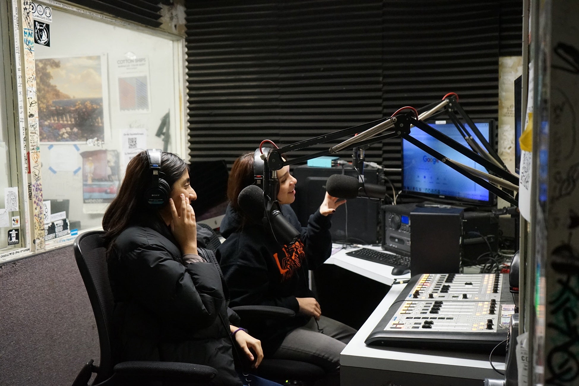 Two women with dark hair sit in front of microphones in radio broadcast booth