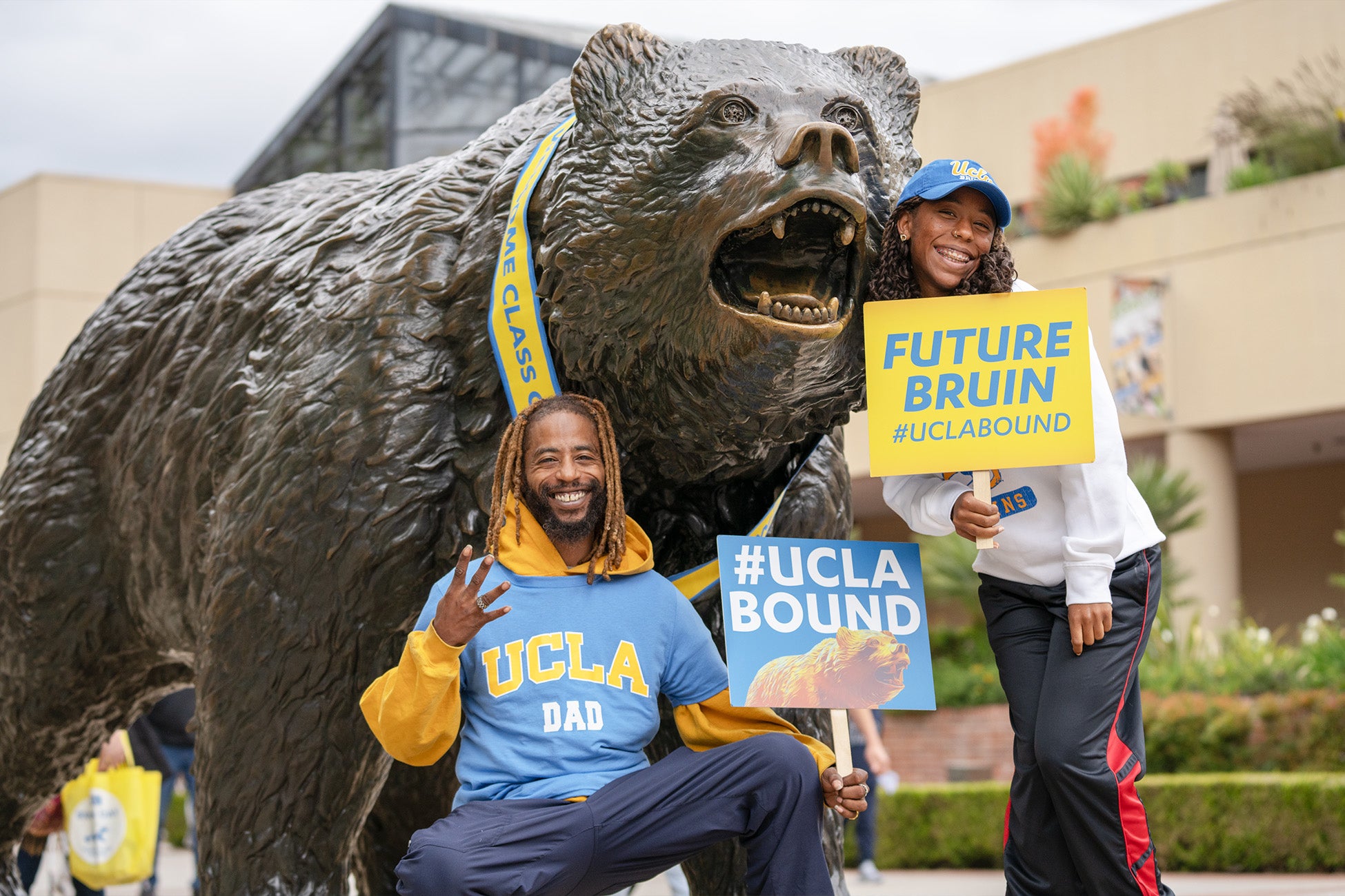 A man and woman pose together with the Bruin Statue, both appearing cheerful, wearing UCLA gear, and holding UCLA bound signs.