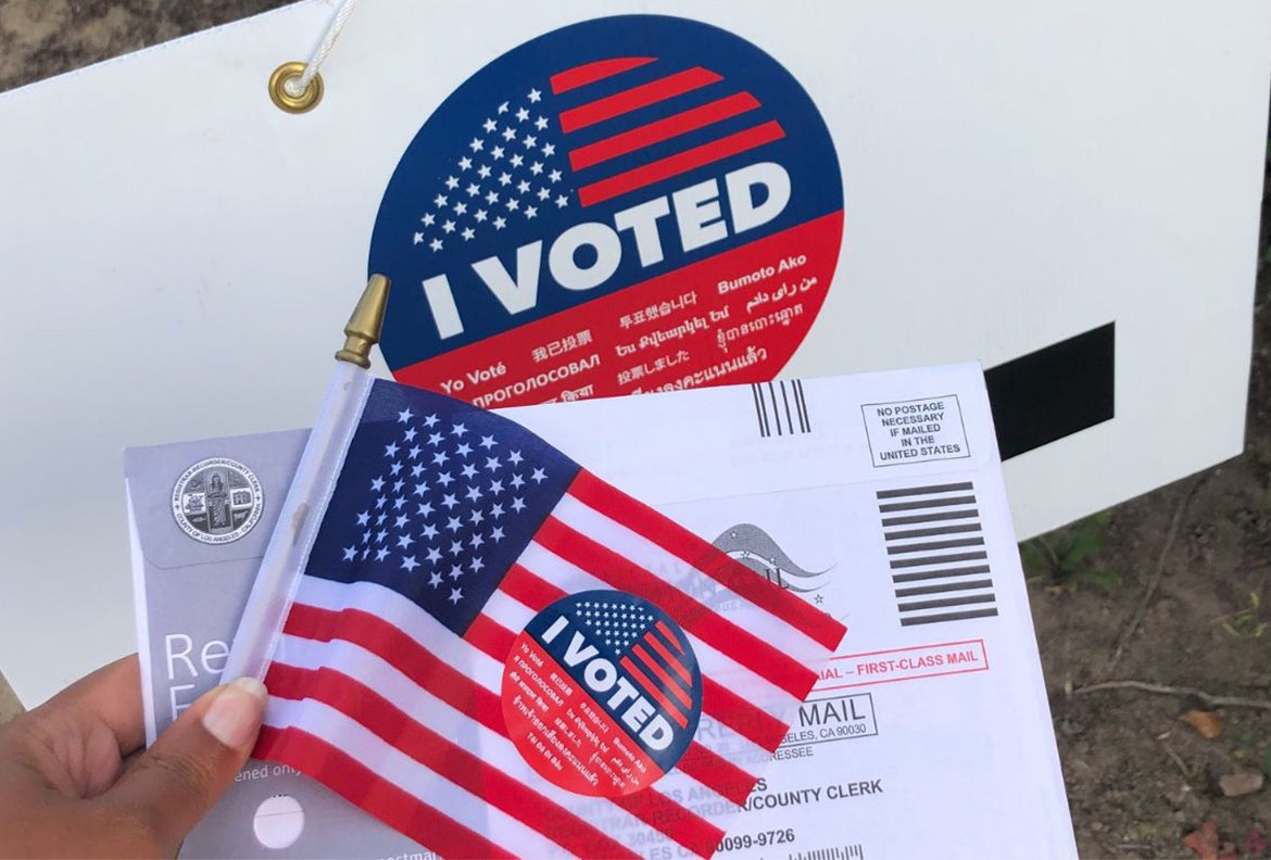 Person holding a small American flag, an ‘I Voted’ sticker and a ballot