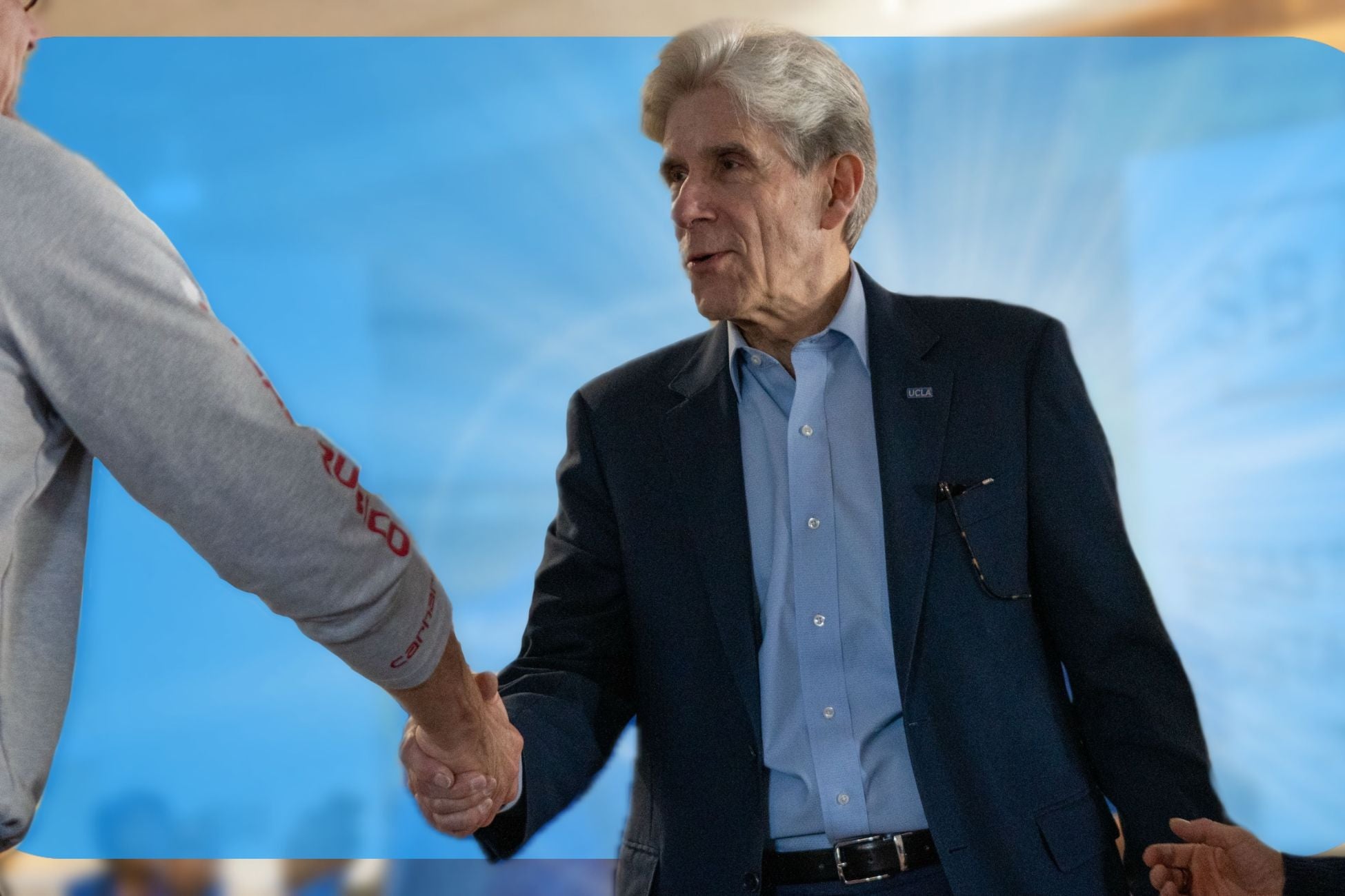 Chancellor Frenk, wearing a suit with UCLA pin, shakes hands with an individual in a gray shirt. 