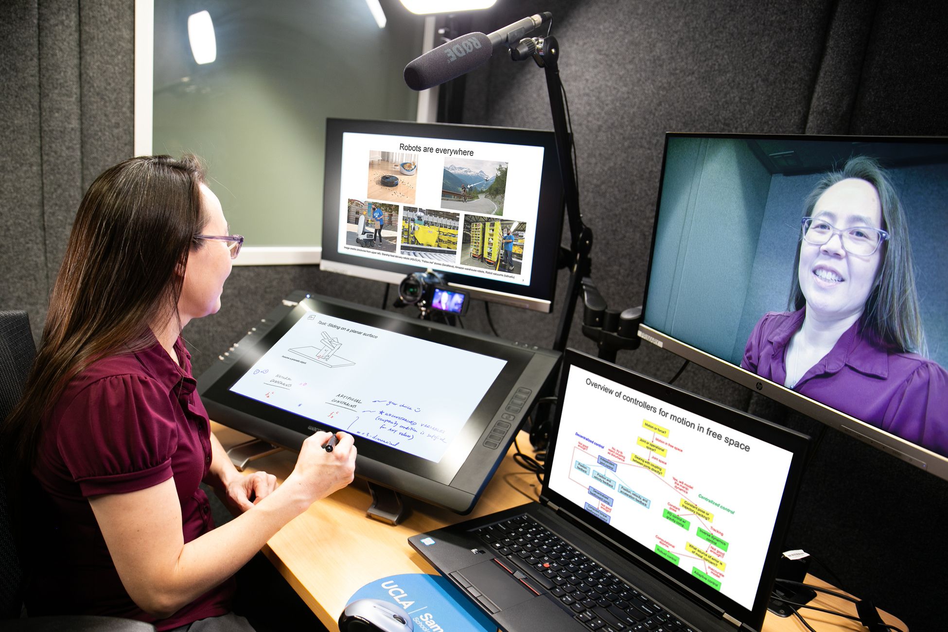 A person sits at a desk using a drawing tablet, communicating through a video call on a monitor. Several screens display presentation slides and information.