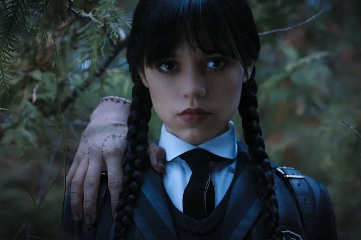 Jenna Ortega in a promo photo from the Netflix series “Wednesday” — dressed in a dark school uniform with braids and a severed hand on her shoulder