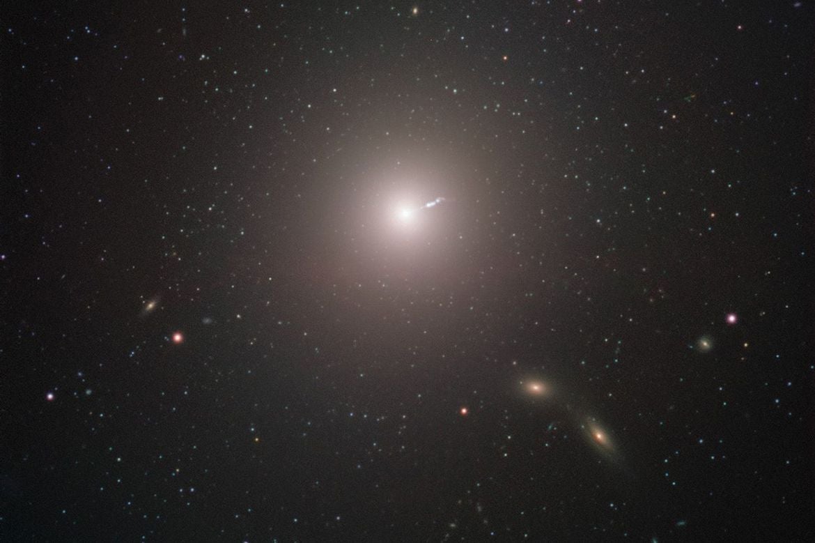 Surrounded by stars and galaxies, the galaxy M87, with a linear “jet” emitting from the center, is seen from a distance as captured by the Event Horizon Telescope