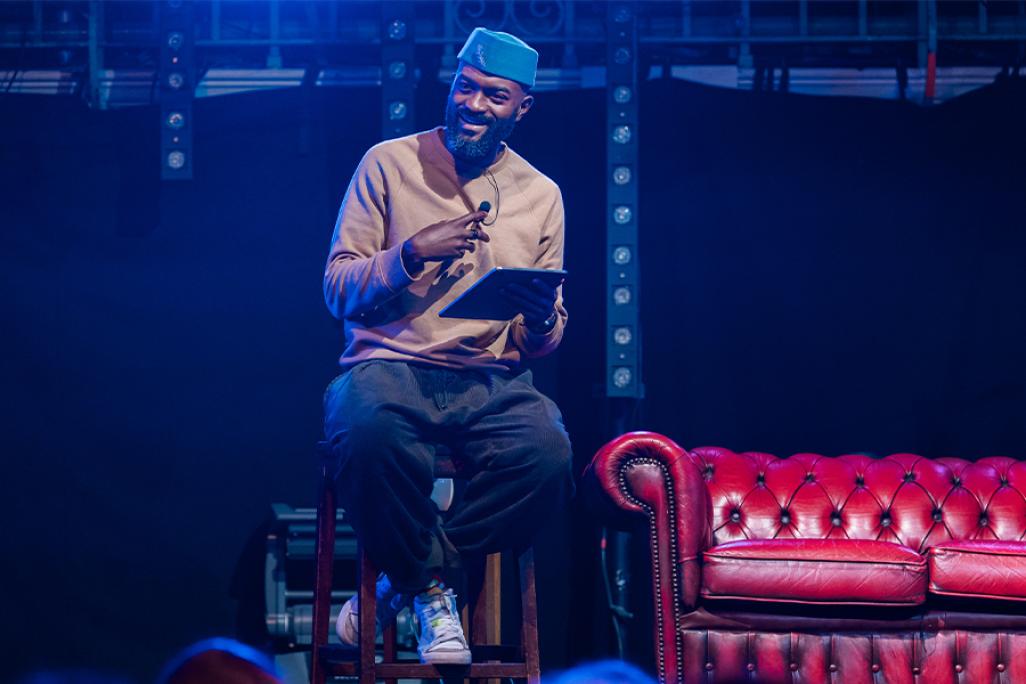 Inua Ellams sitting on stool on stage with mic and tablet