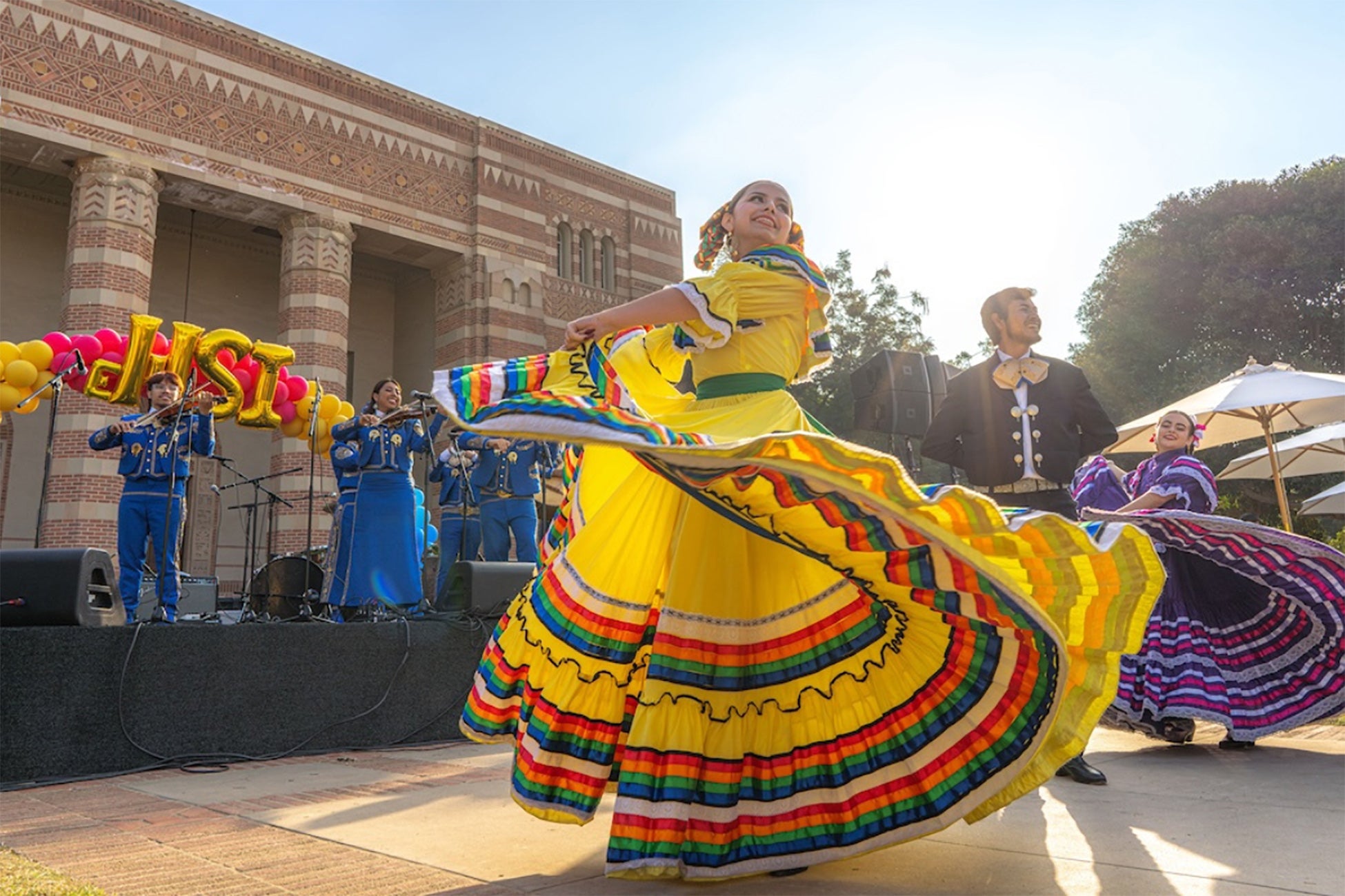 A folklorico dancer in a long yellow dress performs alongside UCLA’s Mariachi de Uclatlán in front of Powell Library.