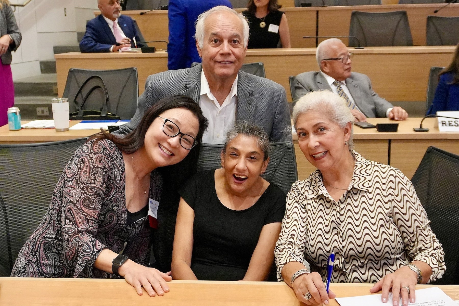 Four people--two sitting, two standing--are behind a desk in an auditorium, with others behind