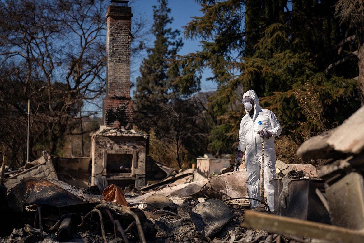 Man in hazardous materials suit picks through ruins of house burned in Altadena