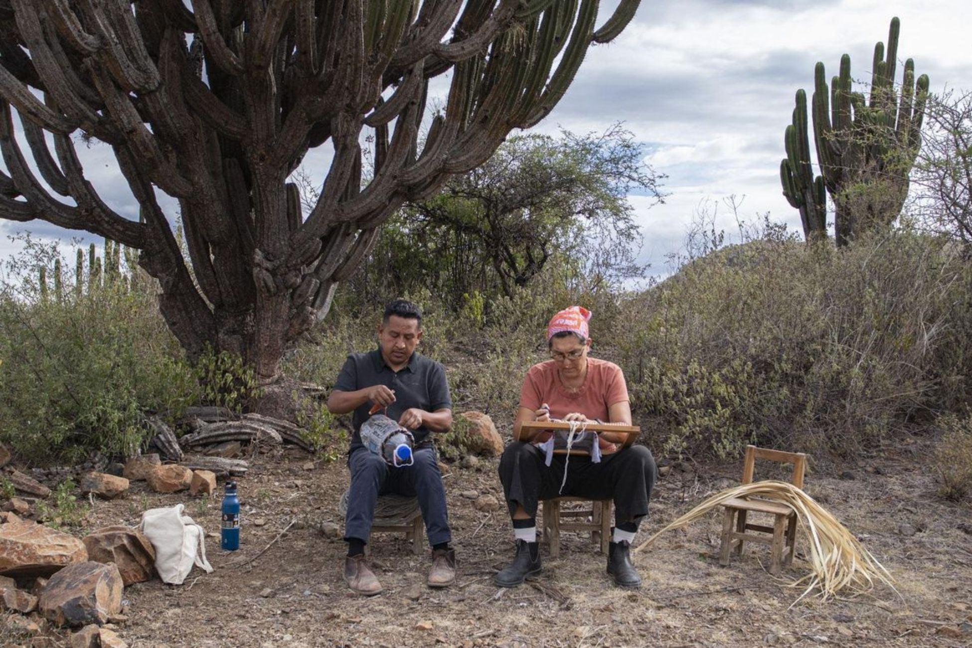 Two people sit on small chairs in a field in Oaxaca, weaving.