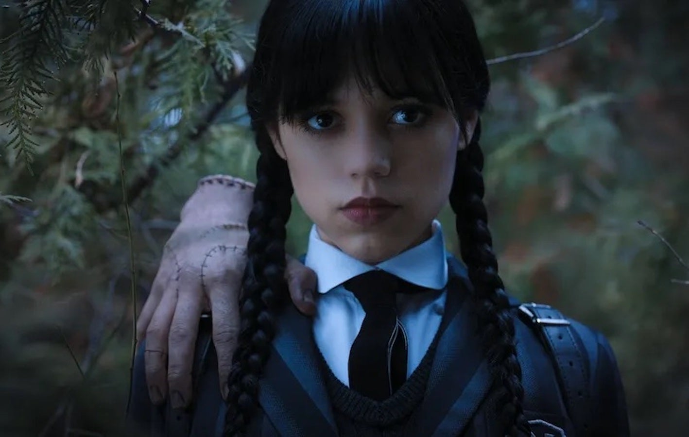 Jenna Ortega in a promo photo from the Netflix series “Wednesday” — dressed in a dark school uniform with braids and a severed hand on her shoulder