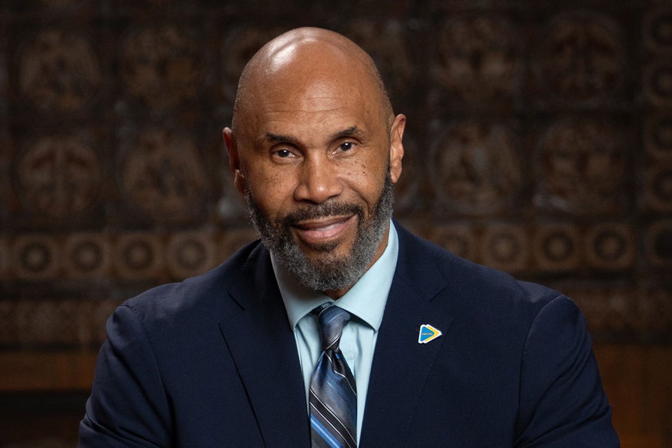 Portrait of a man wearing a navy blue suit, blue striped tie, and a small pin. 