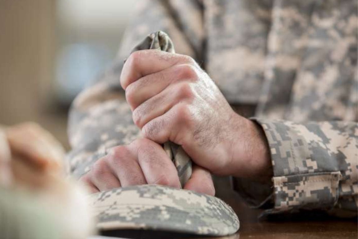 Close-up image shows a man's hands on a desk tightly holding a camouflage hat